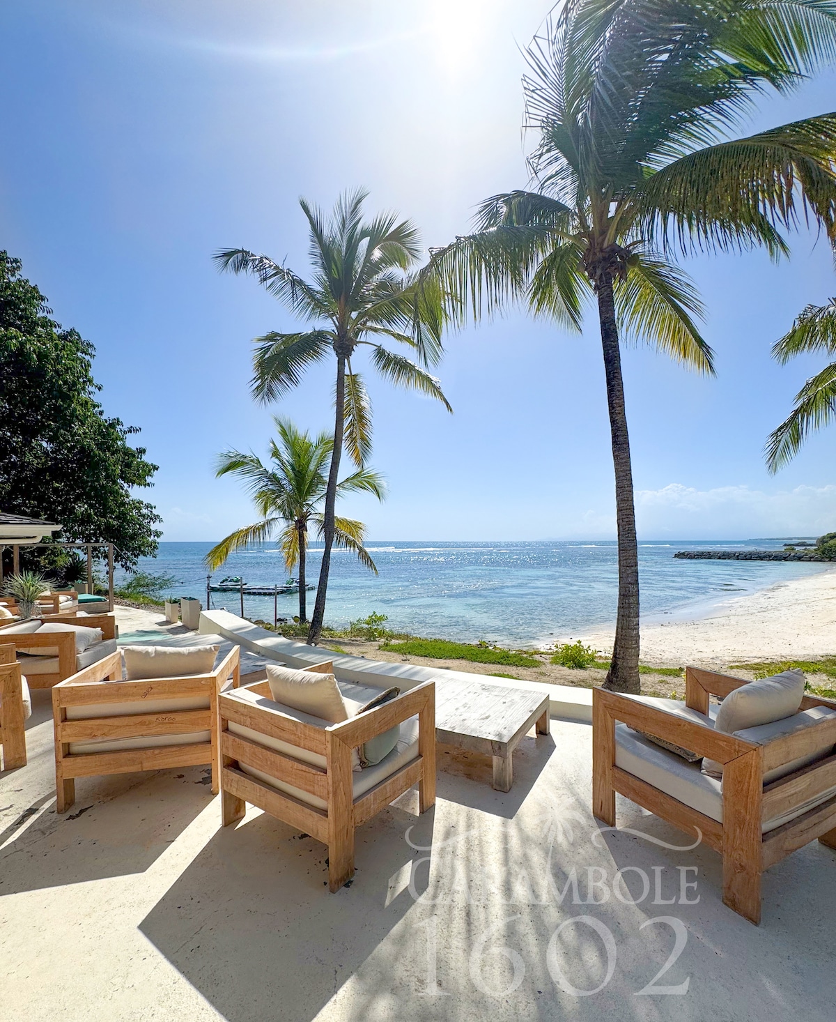 An outdoor seating area is surrounded by palm trees, overlooking the ocean. Wooden lounge chairs with soft cushions provide a comfortable space to relax. The clear blue water and sandy beach are visible in the background, complemented by a bright sky.