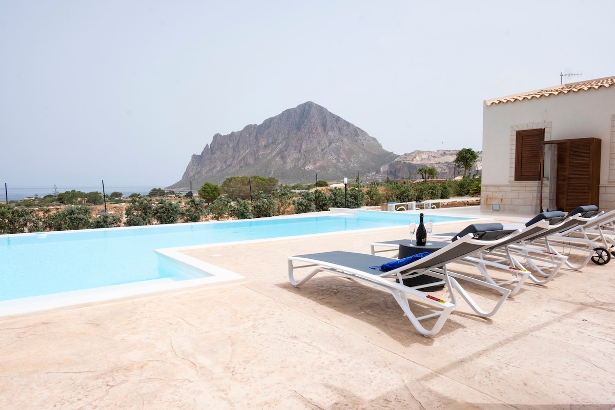 A tranquil scene is presented with a clear swimming pool reflecting the sky. Sun loungers arranged beside the pool allow for relaxation, with a bottle of wine showcased on one. A distant mountain rises in the background, surrounded by greenery.