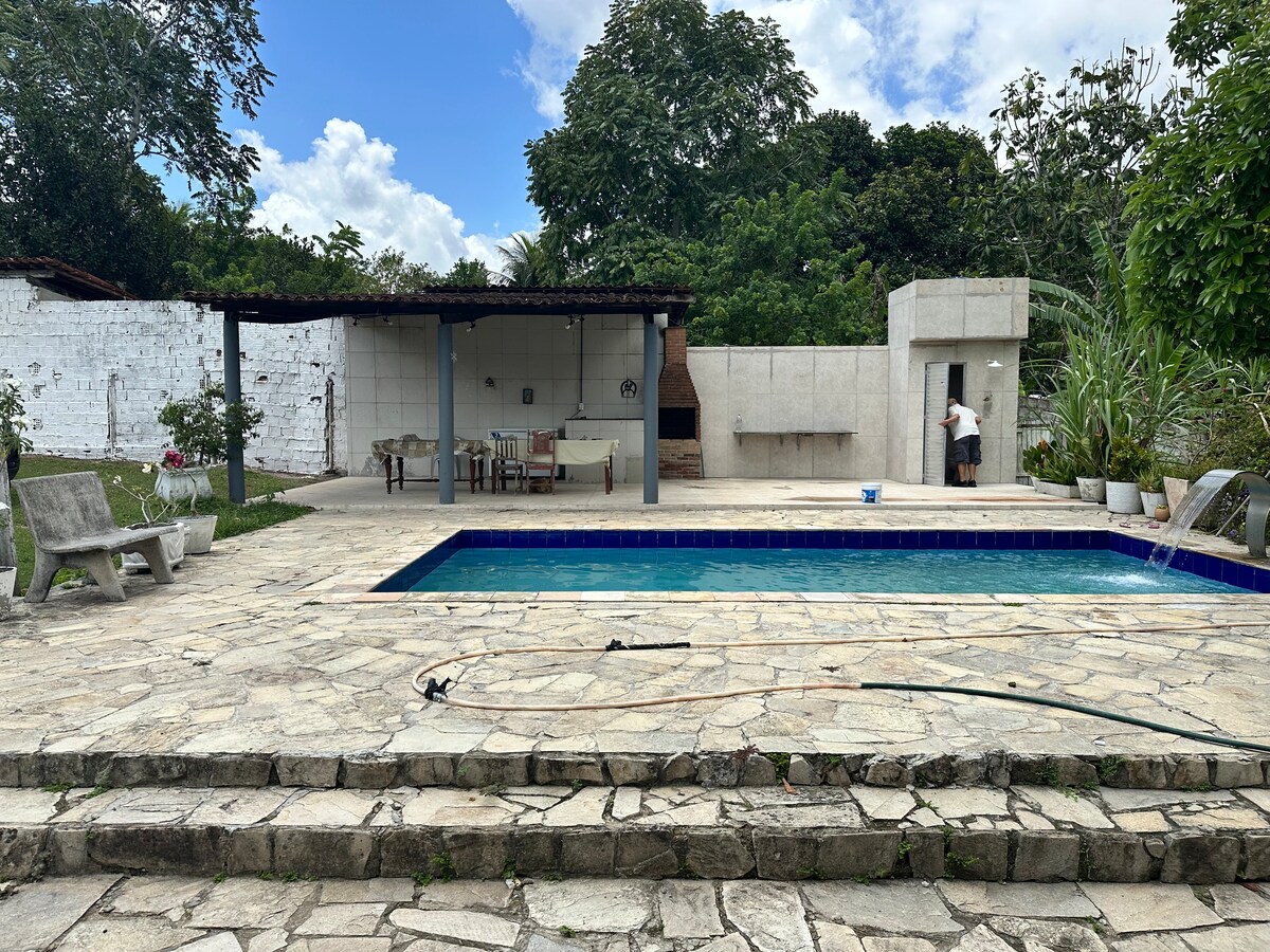 A rectangular swimming pool sits at the center of a spacious outdoor area, surrounded by stone paving. A shaded seating area is visible, featuring a dining table and chairs. Lush greenery surrounds the space, enhancing the natural ambiance.