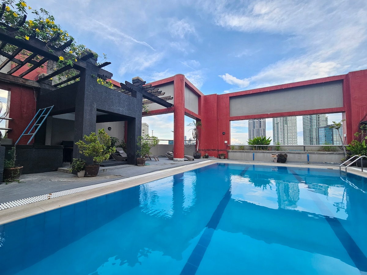 A rooftop pool is shown, featuring clear blue water and surrounded by planters with greenery. Scenic views of city buildings are visible through large open windows, and a shaded seating area is present on one side of the pool.
