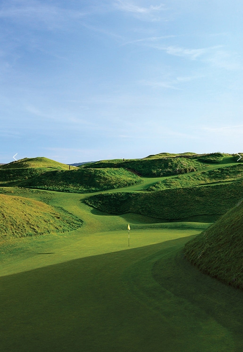 A rolling green landscape is captured, featuring undulating hills covered in lush grass. A golf hole with a flag stands prominently on the manicured fairway, framed by the gentle contours of the terrain. The clear blue sky overhead enhances the vibrant colors of the scene.