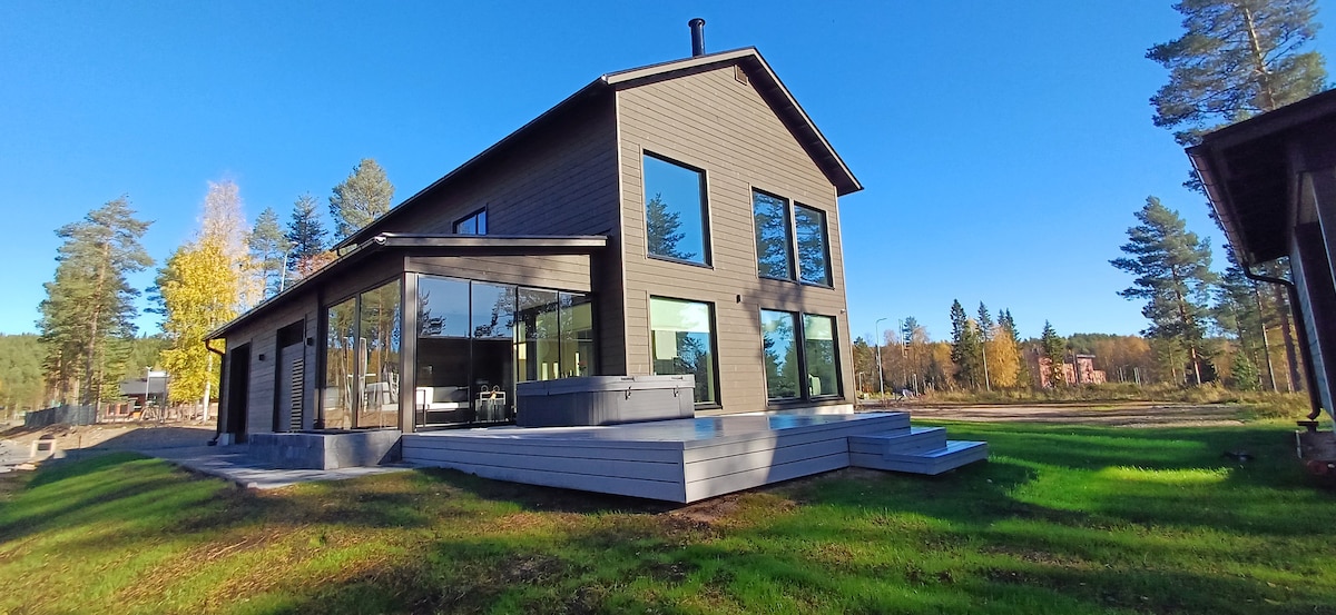 The modern house features large glass windows that enhance natural light and provide views of the surrounding landscape. A hot tub is positioned on the wooden deck, situated in a grassy area with trees in the background, creating a serene outdoor environment.