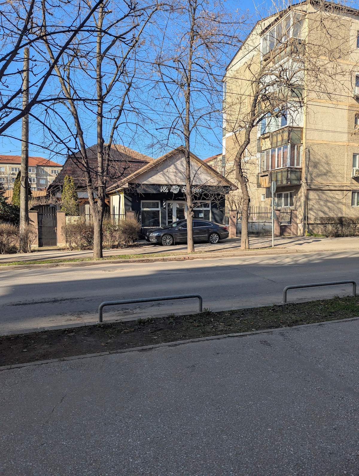 A street view shows a modern building alongside a quaint, single-story structure with a dark roof. Large windows reflect the surroundings. Leafless trees line the street, enhancing the urban landscape. A parked car is visible, contributing to the local atmosphere.
