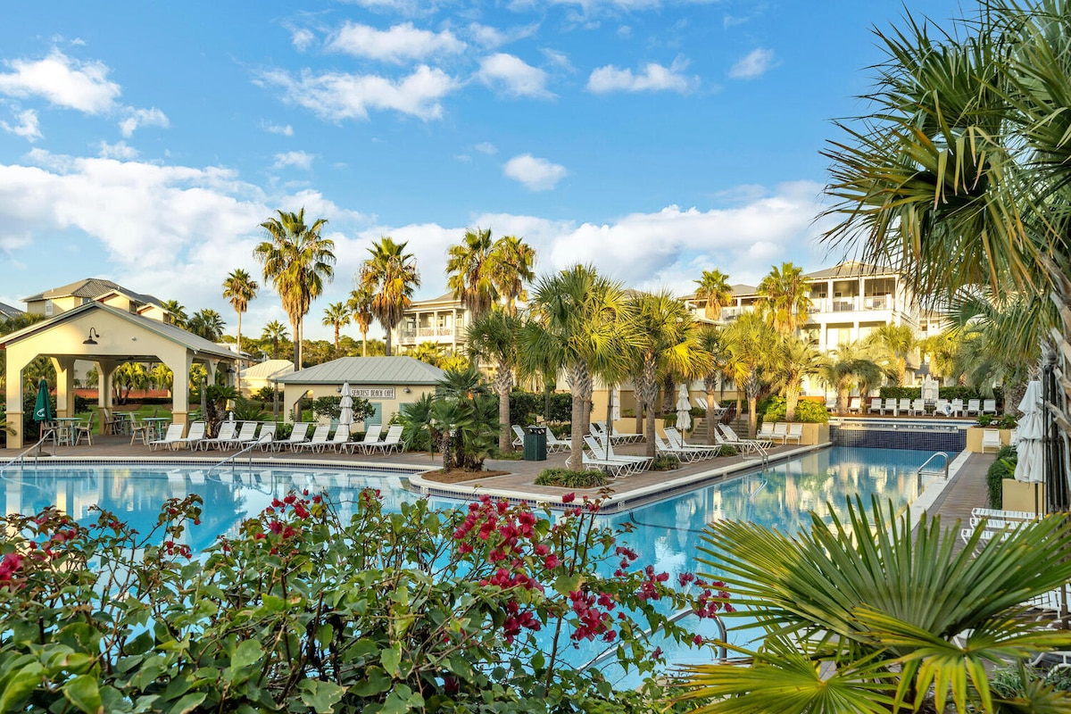 A large lagoon pool is surrounded by lush palm trees and vibrant tropical plants. Sun loungers are positioned along the pool's edge, with a clubhouse visible in the background. Clear blue skies provide a bright setting, enhancing the welcoming atmosphere of the community space.
