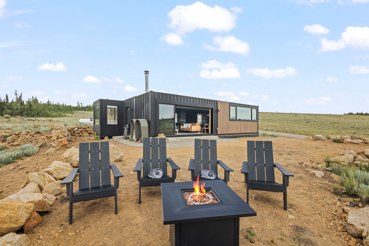 An outdoor seating area features five black chairs arranged around a firepit, offering a space for gatherings. The backdrop includes a modern shipping container home with large windows, set against the expansive Colorado landscape under a clear blue sky.