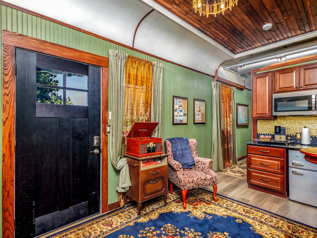 A cozy living area features a plush armchair and a vintage record player, adding a nostalgic touch. The inviting wooden entry door complements the green paneled walls, while framed artwork decorates the space. A patterned area rug enhances the warmth and charm of the room.