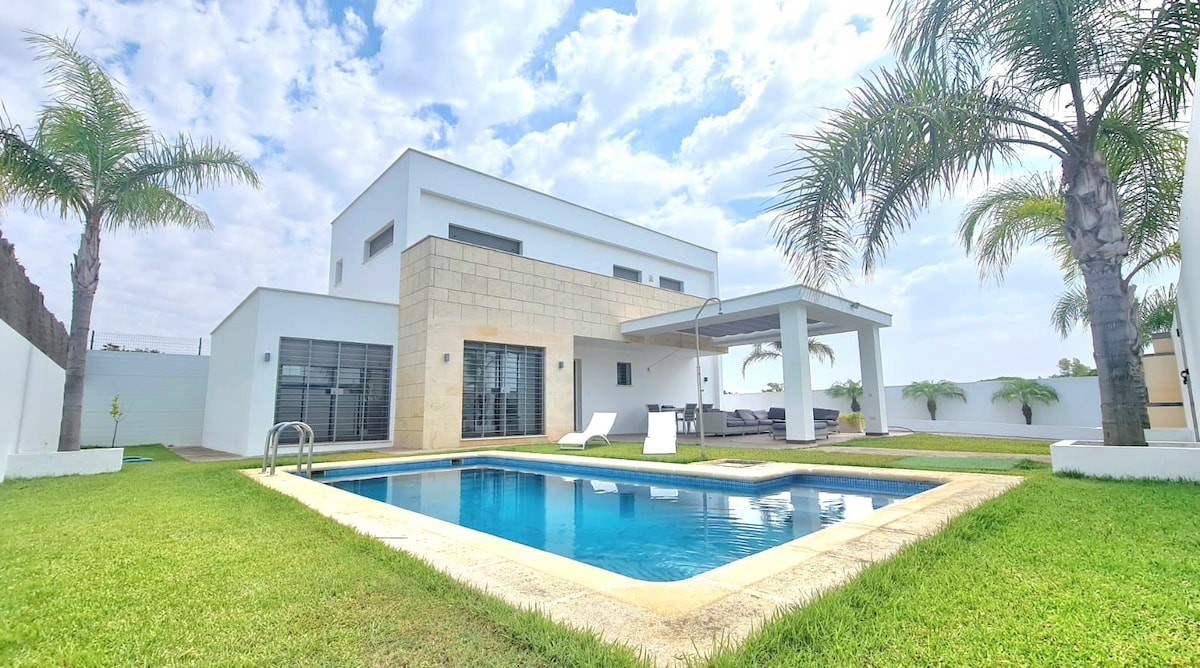 A modern villa is depicted with a spacious pool, surrounded by well-kept grass. Palm trees add softness to the outdoor landscape. A covered patio with lounge chairs offers a shaded area, while large windows provide views into the bright interior.