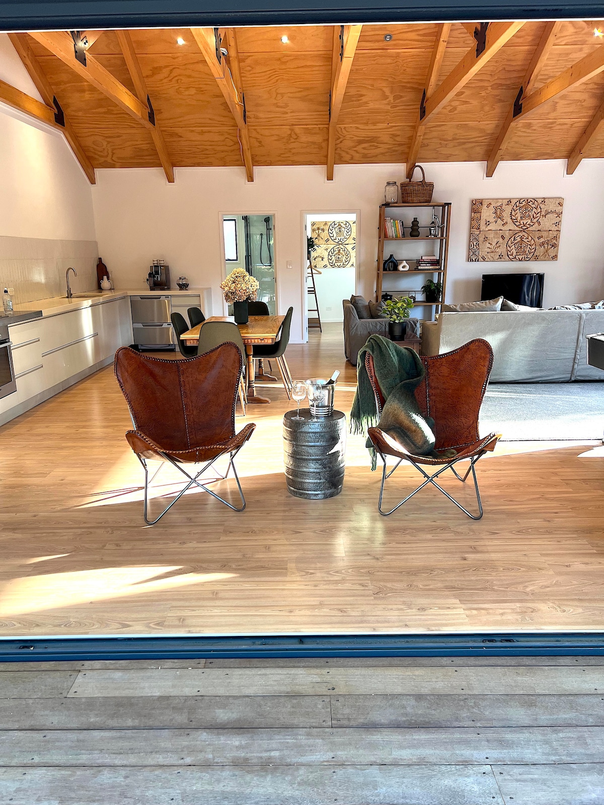A spacious living area is viewed from an open doorway, featuring two leather butterfly chairs in the foreground. Behind them, a dining table is set for four, and a cozy seating arrangement is visible, accented by natural light from large windows and wooden ceiling beams.