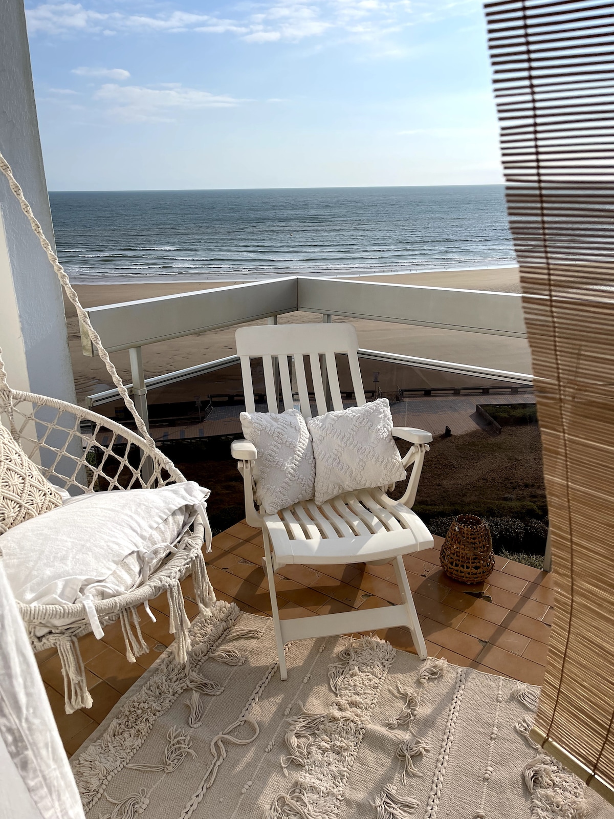 A balcony is featured with a white wooden chair and decorative pillows, overlooking a serene ocean view. A hanging macramé chair adds a cozy touch, while a woven rug complements the space. Natural light enhances the calm atmosphere, framed by light curtains.