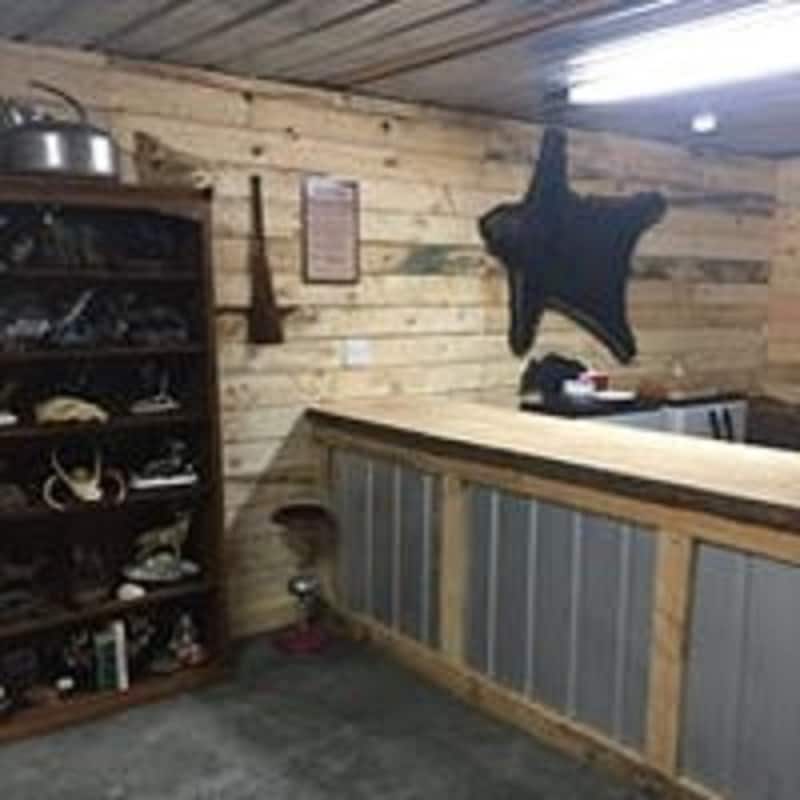 A rustic reception area showcases natural wood paneling and a metal-topped counter. A wooden shelf displays various decorative items, while a mounted star-shaped piece adorns the wall. The space is illuminated by overhead lighting, creating a welcoming environment.