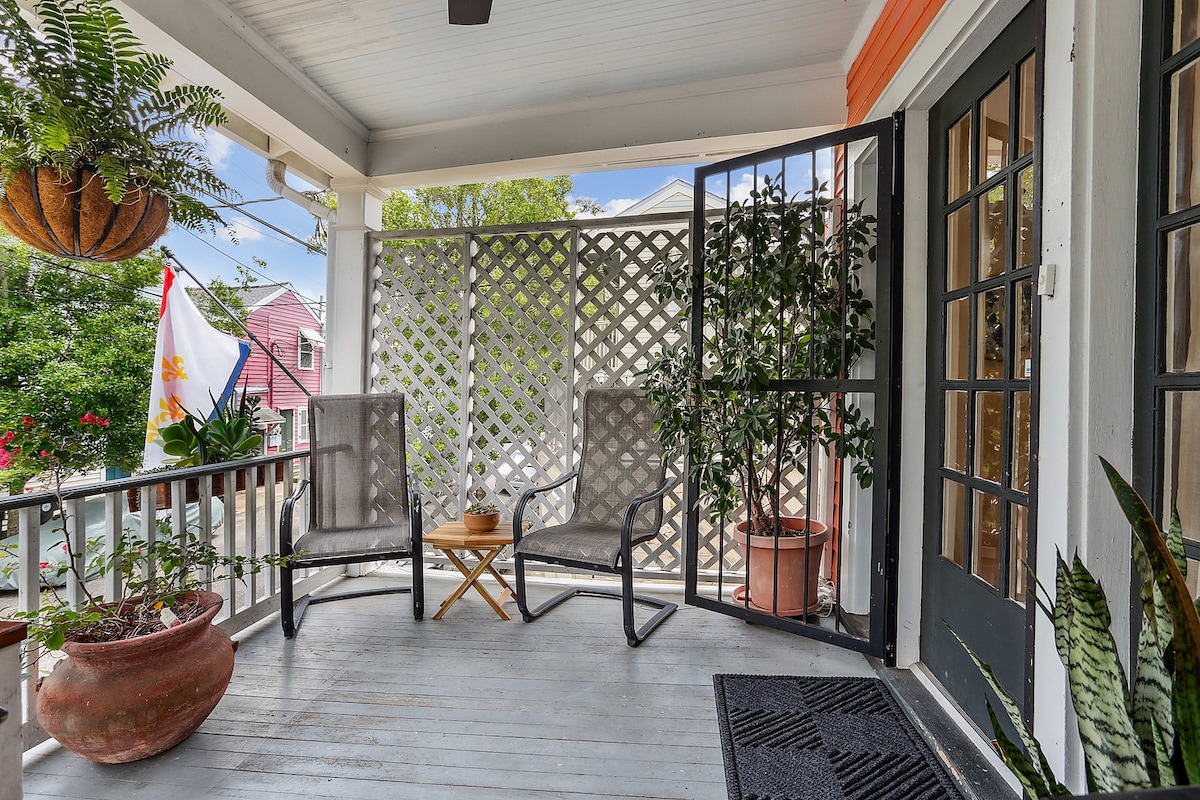 A welcoming porch features two comfortable chairs positioned around a small table, surrounded by greenery from potted plants. A decorative screen provides privacy while a colorful flag adds a touch of charm. Natural light brightens the space, enhancing the inviting atmosphere.