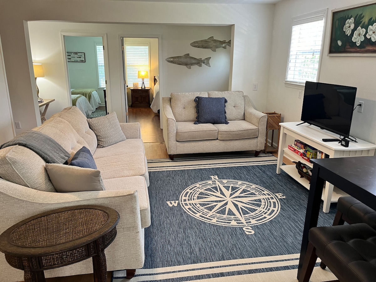 A spacious living area features a light-colored sofa and armchair, arranged around a compass-designed area rug. A wooden table holds games, and a television is positioned atop a stand. Natural light enters through nearby windows, illuminating the soft color palette.