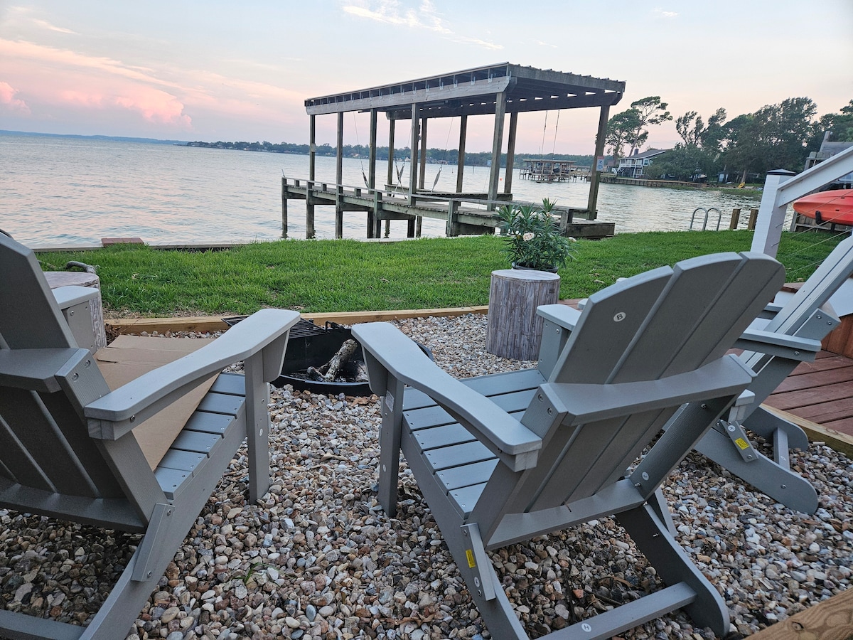 Two gray adirondack chairs are positioned on a stone patio, overlooking Lake Livingston. A wooden dock extends into the water, surrounded by well-maintained grass. A small fire pit is visible nearby, offering a cozy gathering spot.