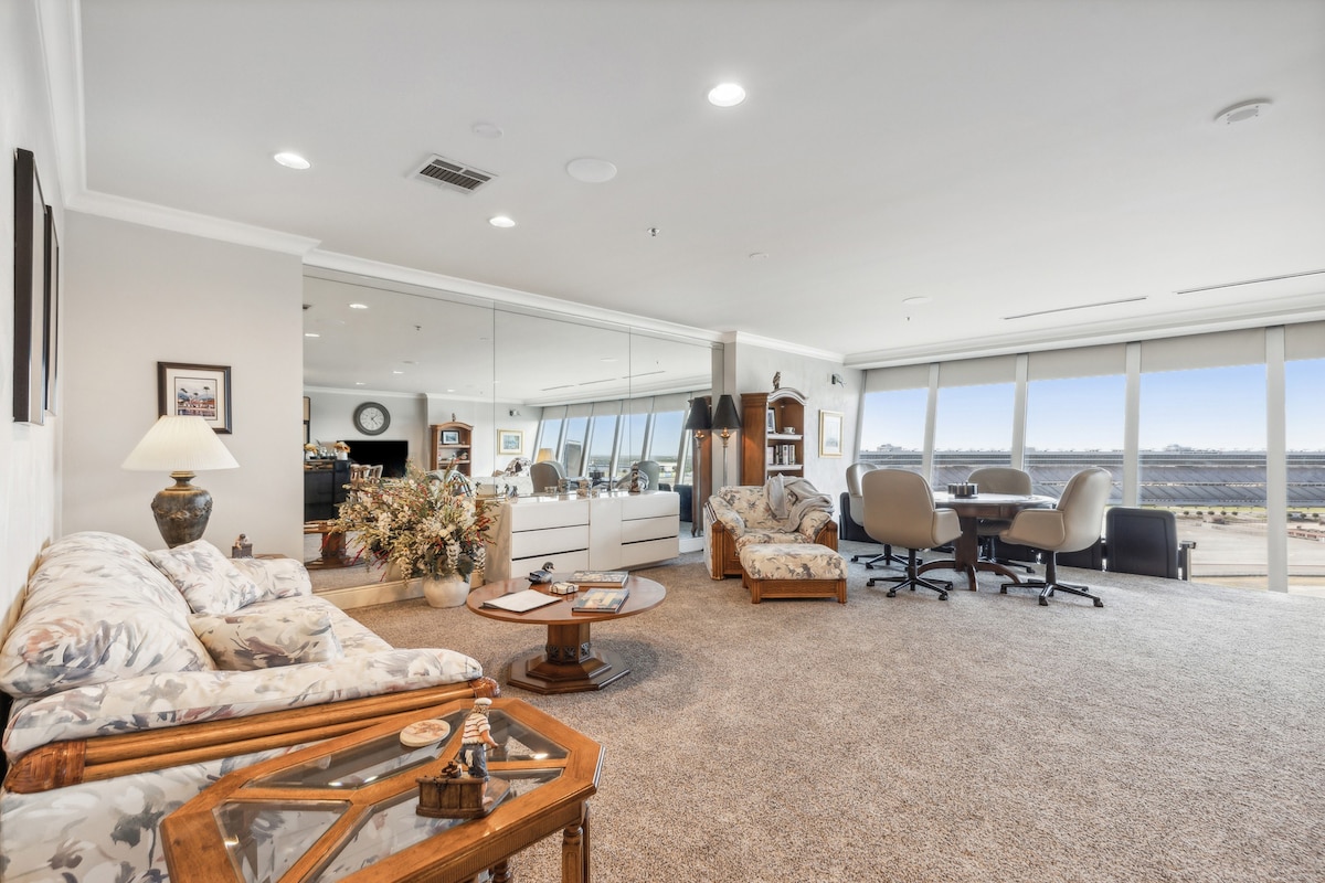A spacious living area is showcased, featuring large windows that offer sweeping views of the racetrack. Two comfortable sofas, adorned with floral patterns, surround a wooden coffee table. A separate dining area with a round table and multiple chairs is also visible, enhancing the inviting atmosphere.