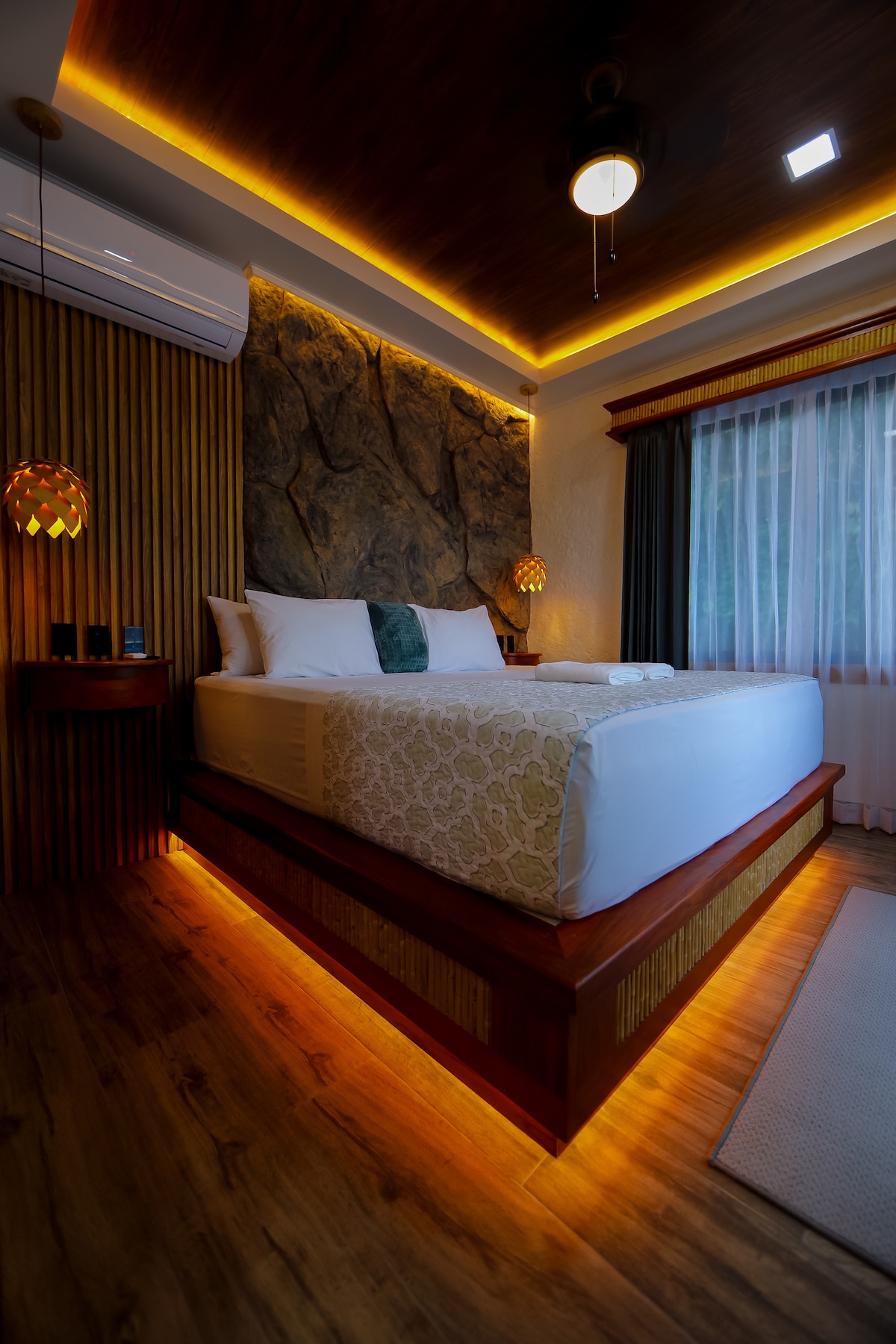 A spacious bed is positioned under a softly illuminated ceiling, enhancing the earthy textures of the wall behind it. Warm lighting highlights the wooden accents and creates a serene atmosphere. Natural light filters through the sheer window treatments, which frame the inviting view outside.