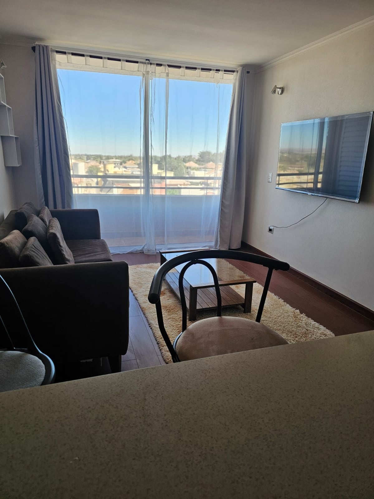 A cozy living area features a comfortable brown sofa set, complemented by a glass coffee table. Large windows adorned with sheer curtains allow natural light to fill the space, with a view of the surrounding area visible outside. A wall-mounted TV adds to the modern ambiance.