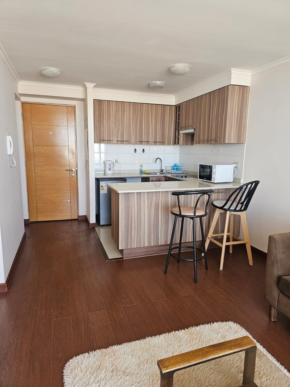 A compact kitchen area is equipped with wooden cabinetry and modern appliances, including a microwave and a refrigerator. Two bar stools are positioned at a sleek counter, while warm wooden flooring complements the neutral-colored walls.