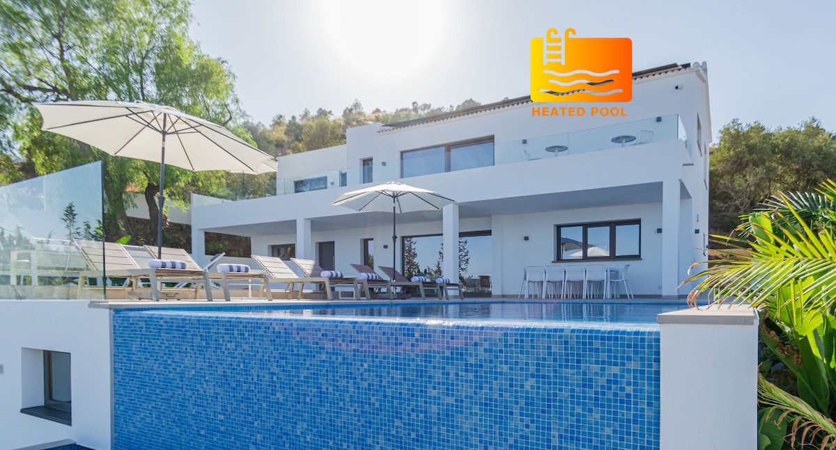 An Ibiza-style villa showcases a stunning heated infinity pool with clear blue tiles. Lounge chairs are arranged under large umbrellas, inviting relaxation. The modern architecture features large windows and a white facade, blending harmoniously with the greenery of the surrounding landscape.