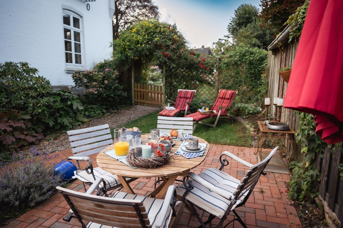 A serene garden space is presented with a wooden table surrounded by four striped chairs. A variety of beverages and pastries are arranged attractively on the table. In the background, two reclining chairs are positioned on lush green grass, bordered by colorful plants and a rustic wooden fence.