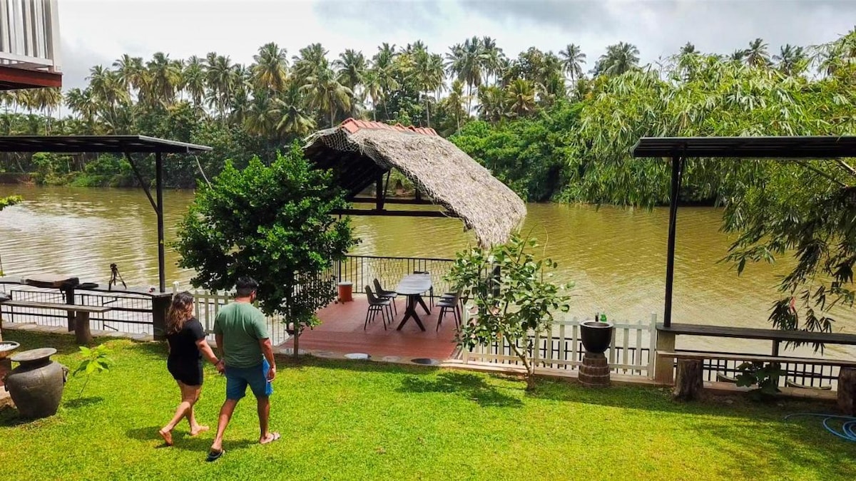 A scenic view of a grassy area leading to the riverbank, with a thatched-roof gazebo overlooking the water. A small patio with a table and chairs is also visible. Lush greenery surrounds the property, enhancing the serene riverside setting.