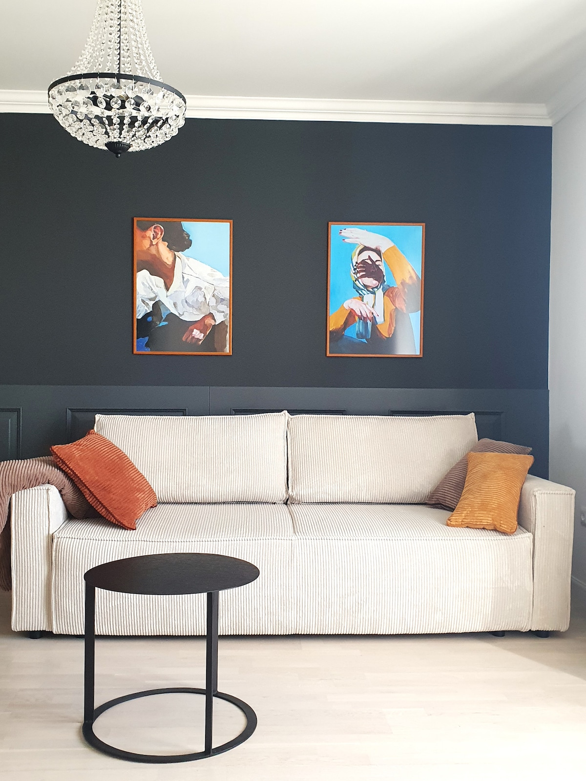 A neutral-toned sofa is positioned in front of a dark blue wall, adorned with two framed artworks. A round black side table sits nearby, complemented by decorative pillows in orange hues. A stylish chandelier hangs above, adding an elegant touch to the space.