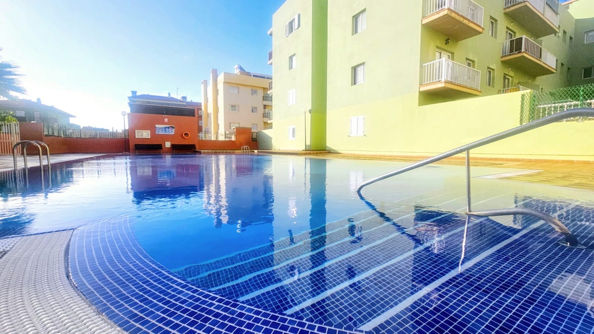 A spacious pool area is captured, featuring clear blue water reflecting sunlight. Surrounding buildings are visible, providing a vibrant backdrop with a mix of colors. Steps lead into the pool, inviting relaxation. The area is well-maintained and offers a serene atmosphere.