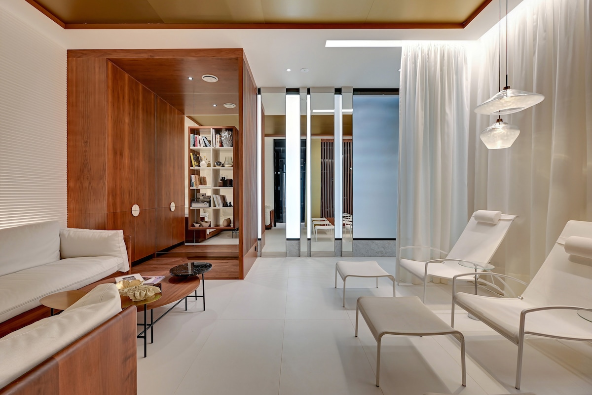 A modern lounge area features sleek lounge chairs positioned around a wooden coffee table. Warm wood accents provide a cozy contrast to the bright white flooring. Floor-to-ceiling curtains gently separate spaces, while built-in shelving displays an array of books in the background.