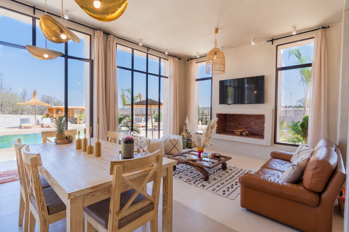 The spacious living and dining area features large windows allowing ample natural light. A wooden dining table accommodates multiple guests, while a comfortable leather sofa is arranged nearby. A modern fireplace and a flat-screen TV add to the cozy atmosphere, complemented by tasteful decorative elements.