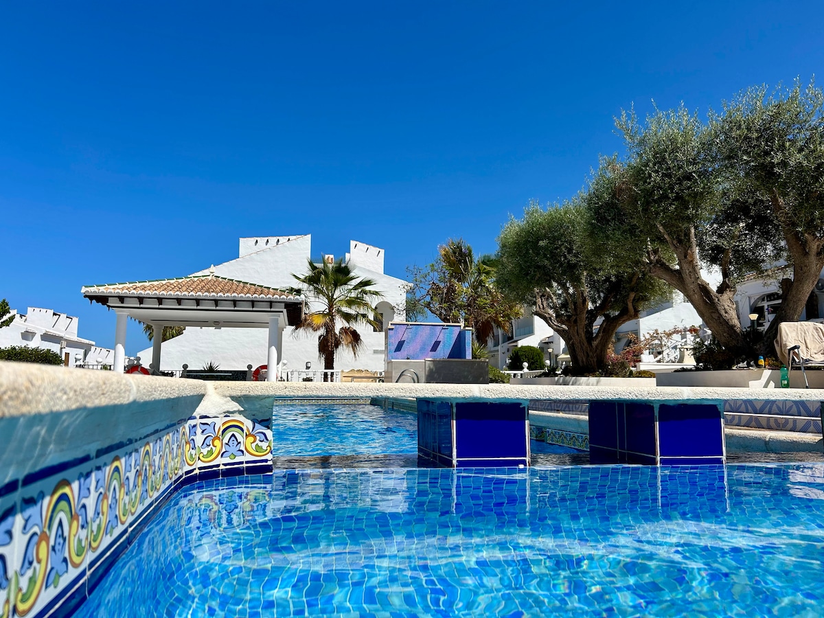 A serene setting is depicted with a sparkling blue pool, framed by tiled edges. Lush trees and a spacious terrace are visible in the background, alongside a white building reflecting the sunny sky. The clear water invites relaxation and enjoyment.