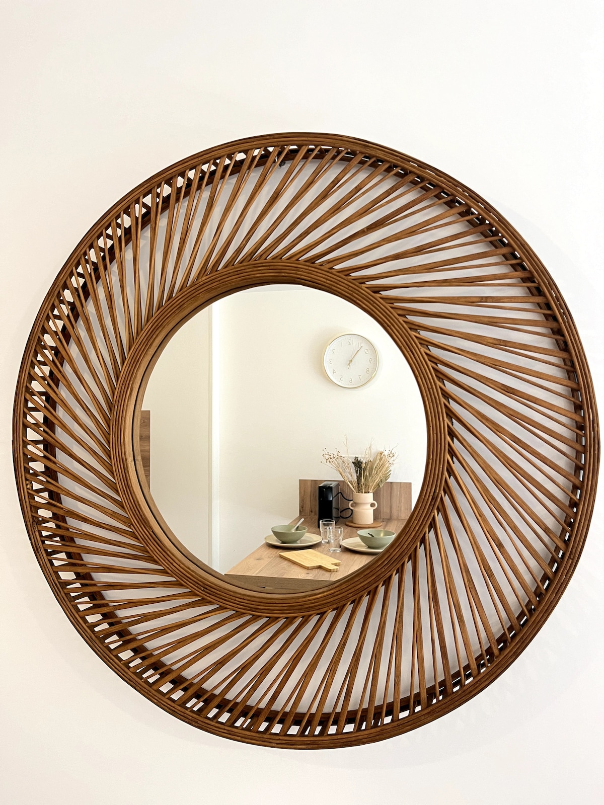 A round mirror with a woven wooden frame features a sunburst design. The reflection includes a dining table set with plates and a small vase, while a wall clock adds to the decor on the white wall behind.