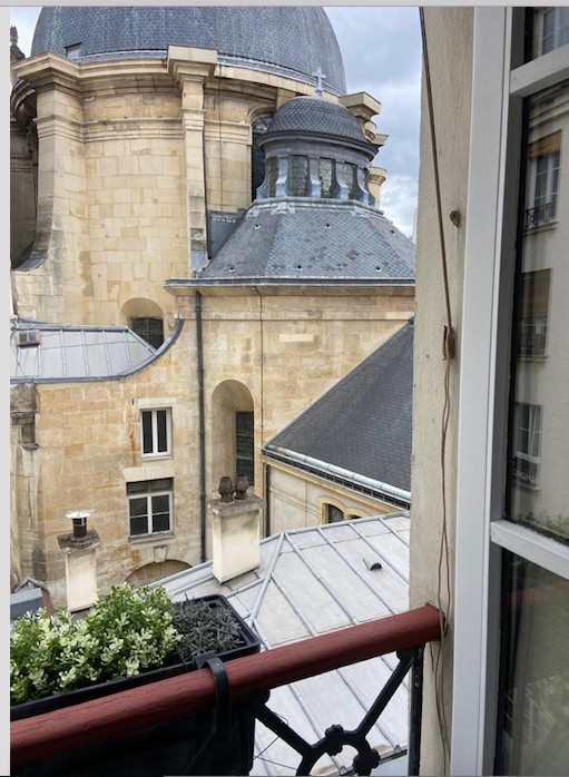 A view from the balcony reveals traditional architectural details of a church with a prominent dome and ornate stonework. The scene is framed by a planter with greenery, and rooftops in various shades of gray are visible under a cloudy sky.