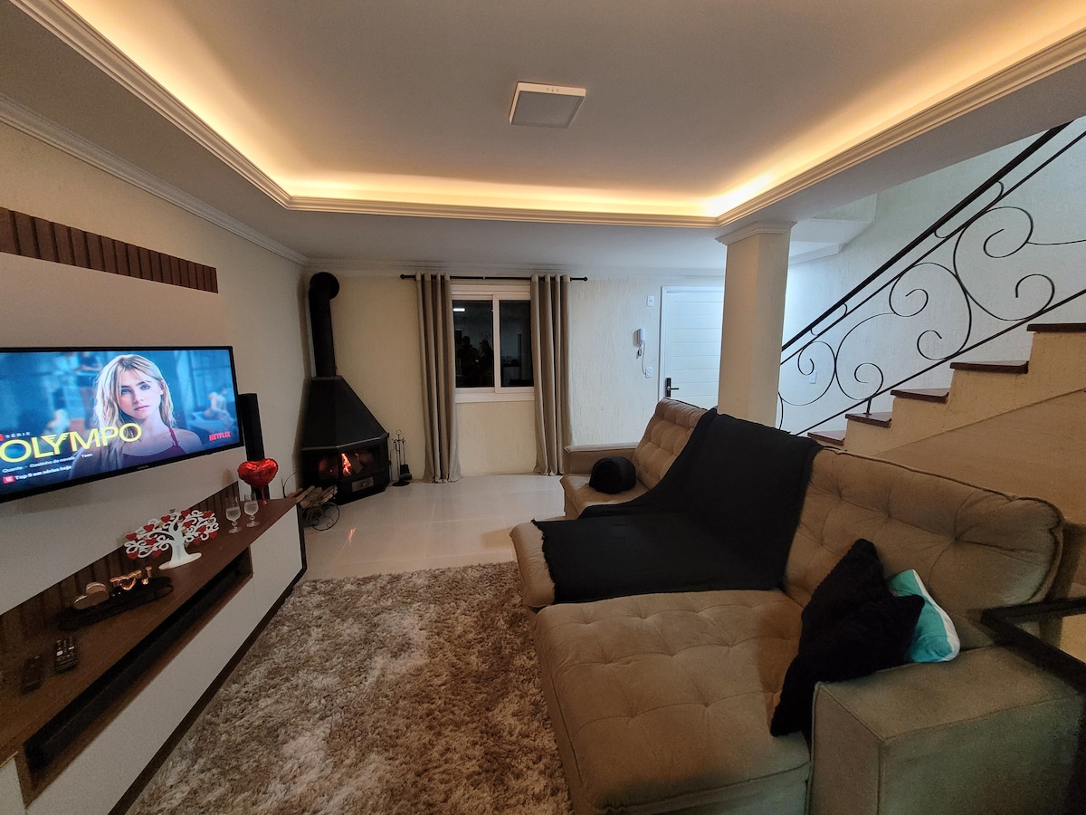A modern living room features a plush, light-colored sectional sofa with dark accents. A wall-mounted Smart TV displays a vibrant image. A black fireplace adds warmth. Soft lighting highlights the space, while a staircase with ornate railings leads to an upper level.