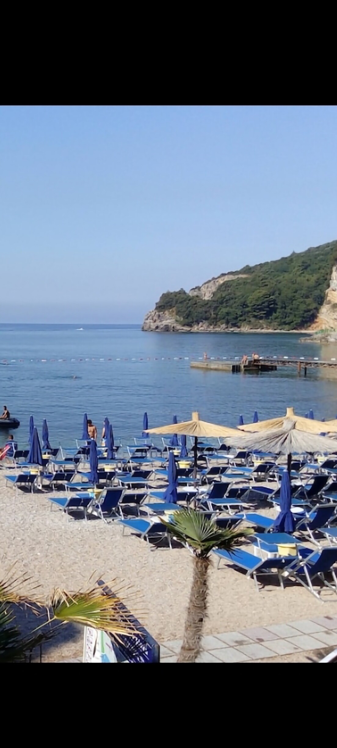 A sandy beach is filled with rows of blue lounge chairs and umbrellas, gently facing the calm sea. The shoreline is lined with greenery and rocky cliffs in the background, while a small dock extends into the water, adding to the coastal atmosphere.