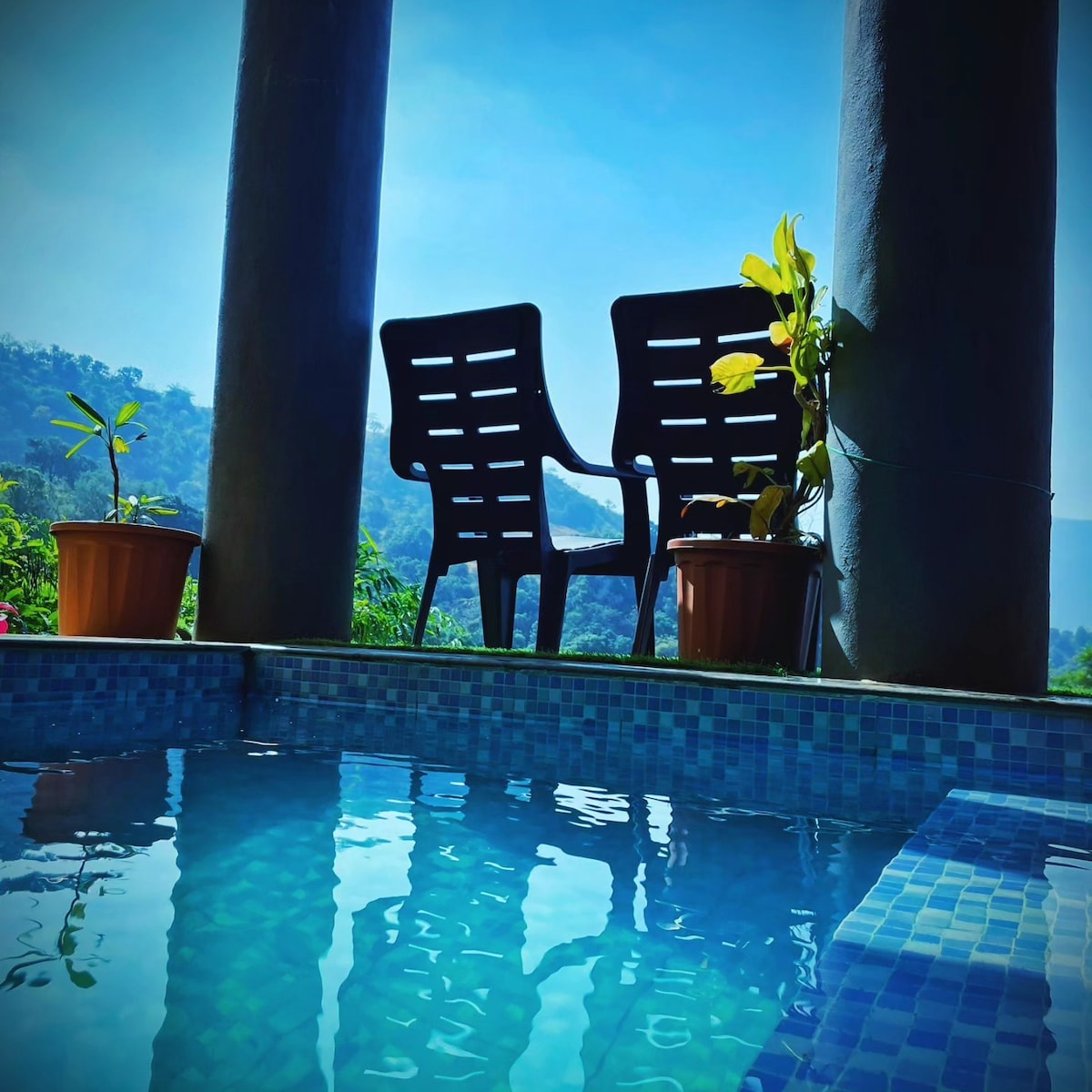 Two black chairs are positioned beside a tranquil plunge pool, with potted plants on either side. The vibrant blue water reflects the clear sky, and gentle hills can be seen in the background, providing a serene environment.