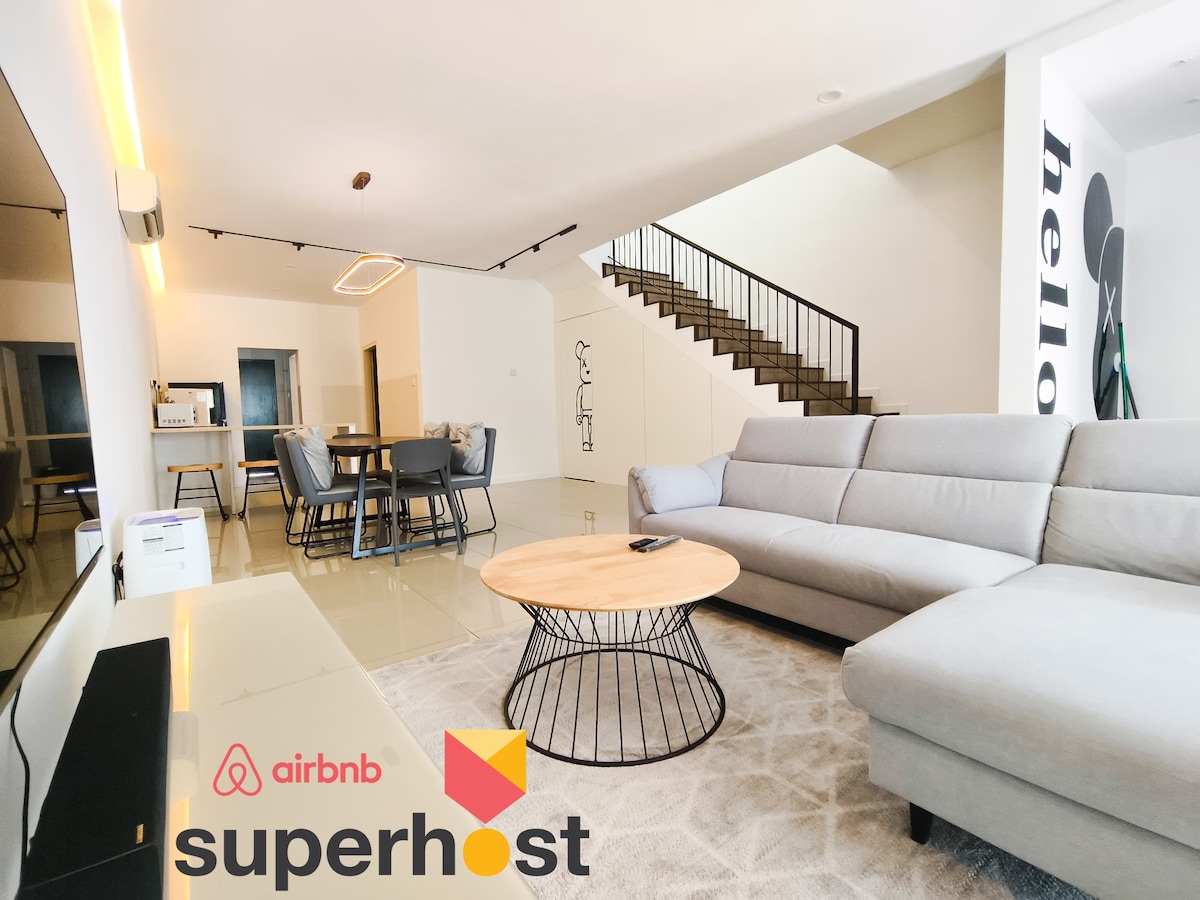 A spacious living area features a light-colored sectional sofa and a round wooden coffee table, complemented by a flat-screen TV on a white console. A dining area with a table for six is visible, along with a staircase leading to an upper level.