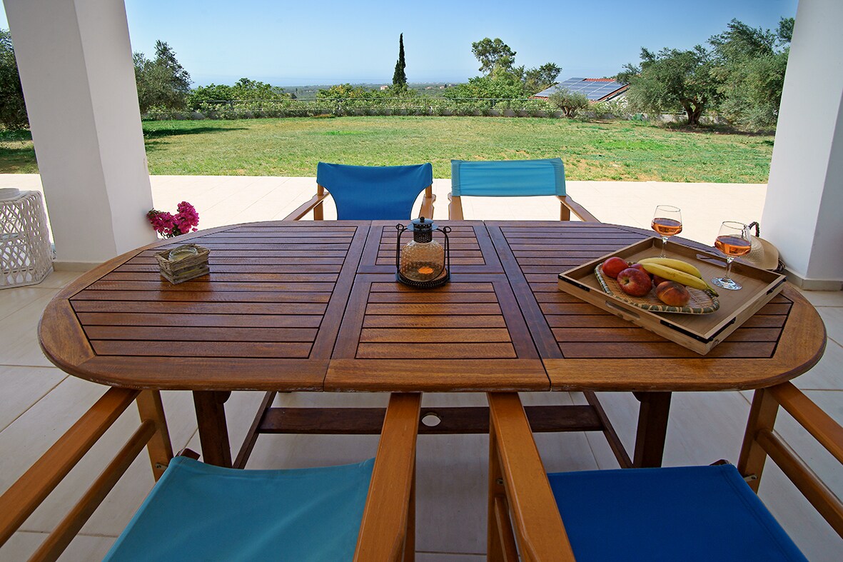 An outdoor dining table made of warm wood is set with a platter of fresh fruits and drinks. Four blue chairs surround the table, while a tranquil garden landscape stretches into the distance, offering views of green trees and the sea.