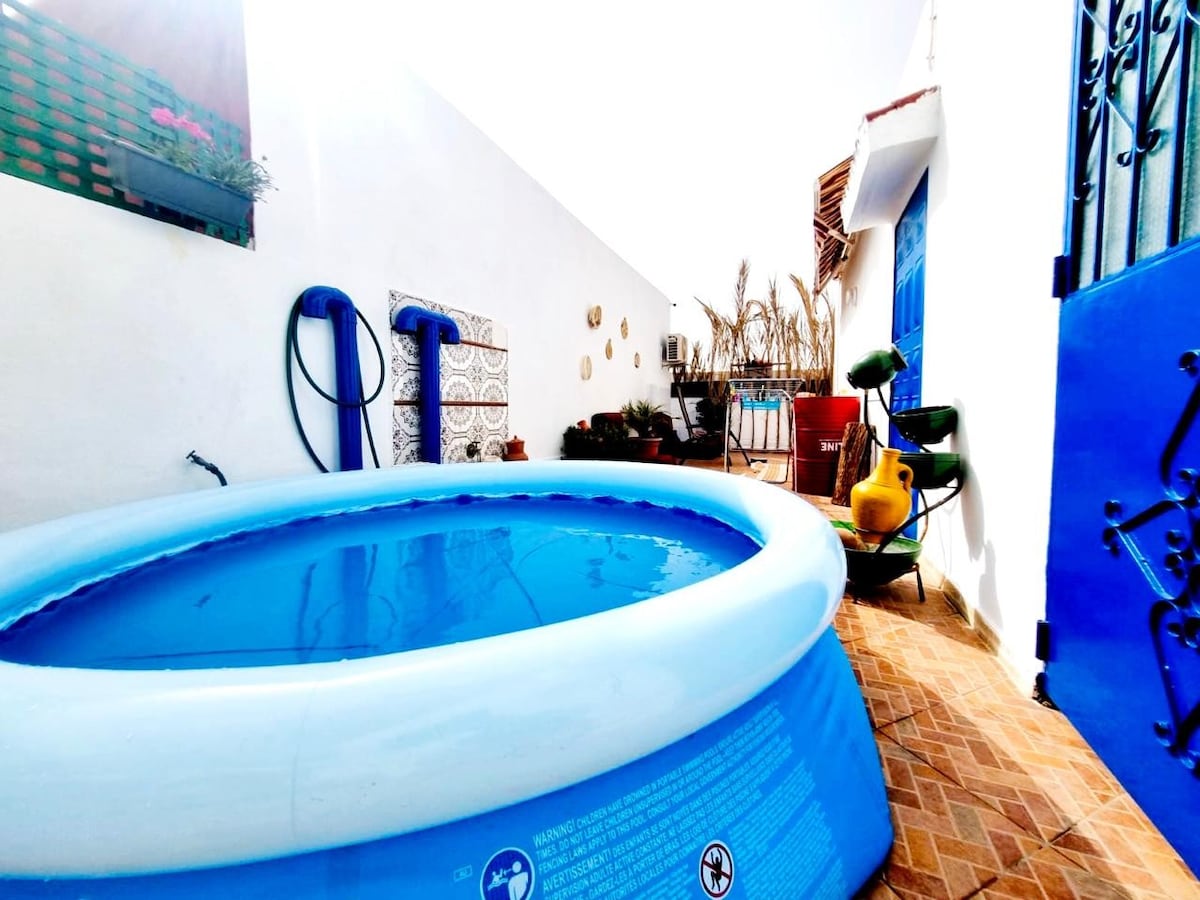 A private terrace is featured, showcasing a round inflatable pool surrounded by tiled flooring. Vibrant blue walls complement various potted plants and outdoor seating arrangements, enhancing the space for relaxation. The area is well-lit, suggesting a bright and inviting atmosphere.