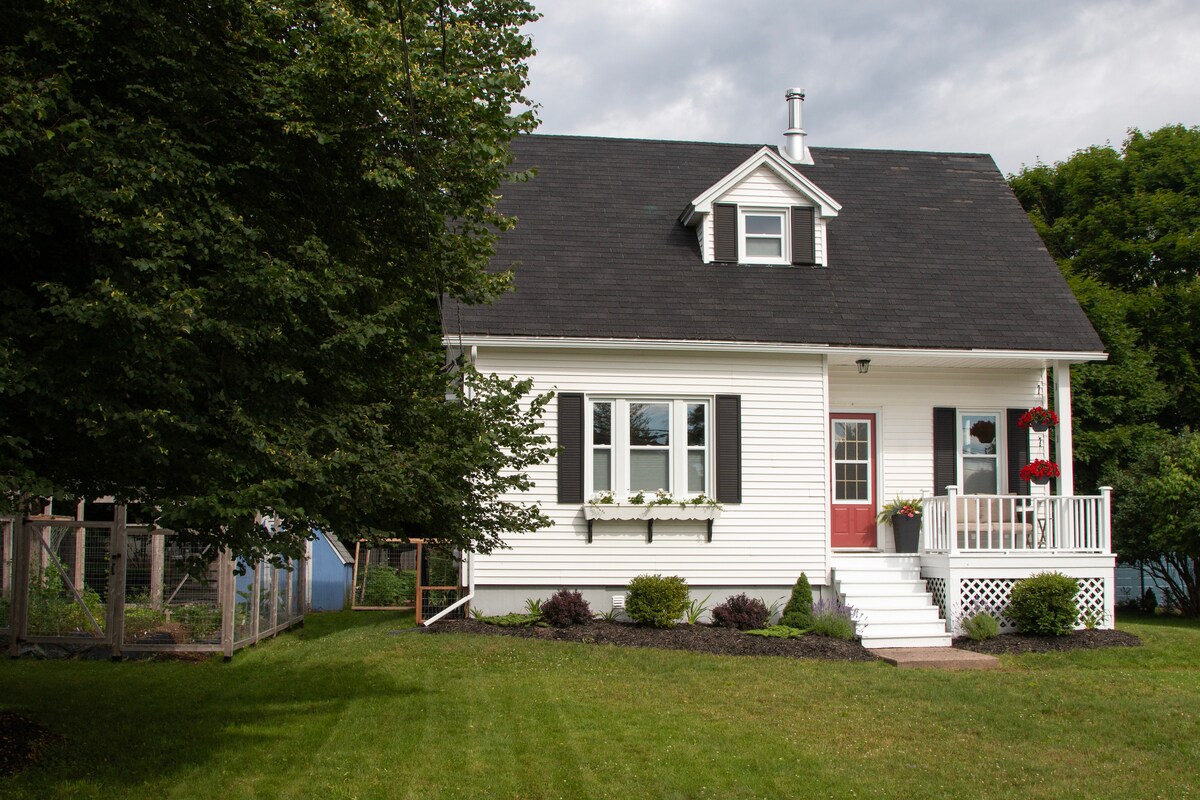 A charming two-story home with white exterior and black shutters is nestled in a lush grassy yard. Flower boxes adorn the windows, adding a touch of color. A small porch with railings invites relaxation, while a garden area can be seen in the background.