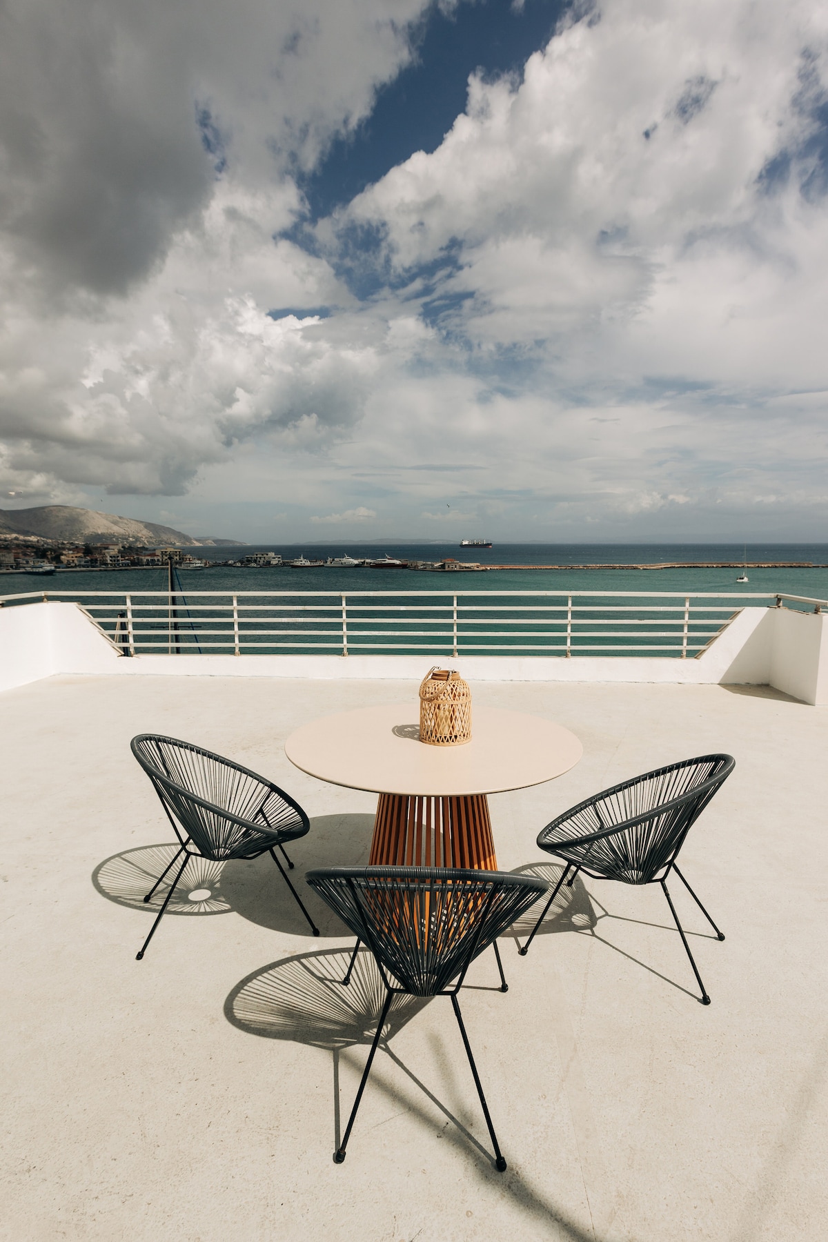 A spacious terrace is featured with a round table and four woven chairs, positioned to overlook the harbor. The surface is smooth and light-colored, while the sky above showcases dynamic clouds. Views of the water and surrounding landscape are prominently visible.