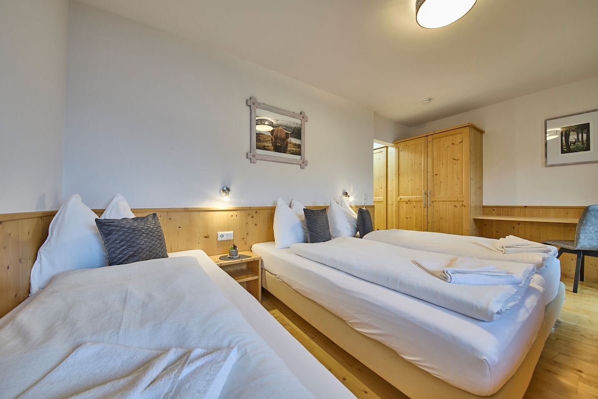 A spacious guest room features two neatly arranged beds with soft linens. Natural wood accents are present in the furnishings, including a wardrobe and a chair. Soft lighting illuminates the room, creating a calming atmosphere. A decorative wall picture enhances the overall decor.