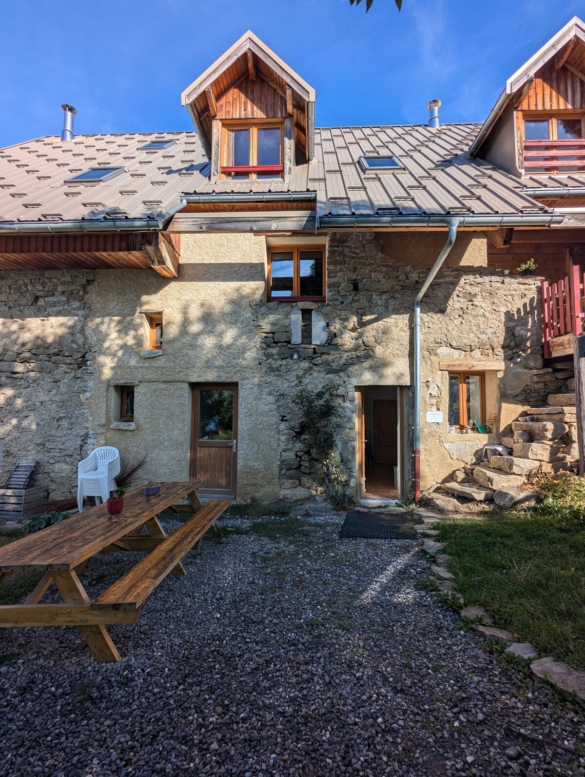 The exterior of a charming stone building is featured, showcasing a sloped roof and multiple windows. A wooden picnic table is set on a gravel area, surrounded by greenery. The entrance is visible, along with a staircase leading to a higher level.
