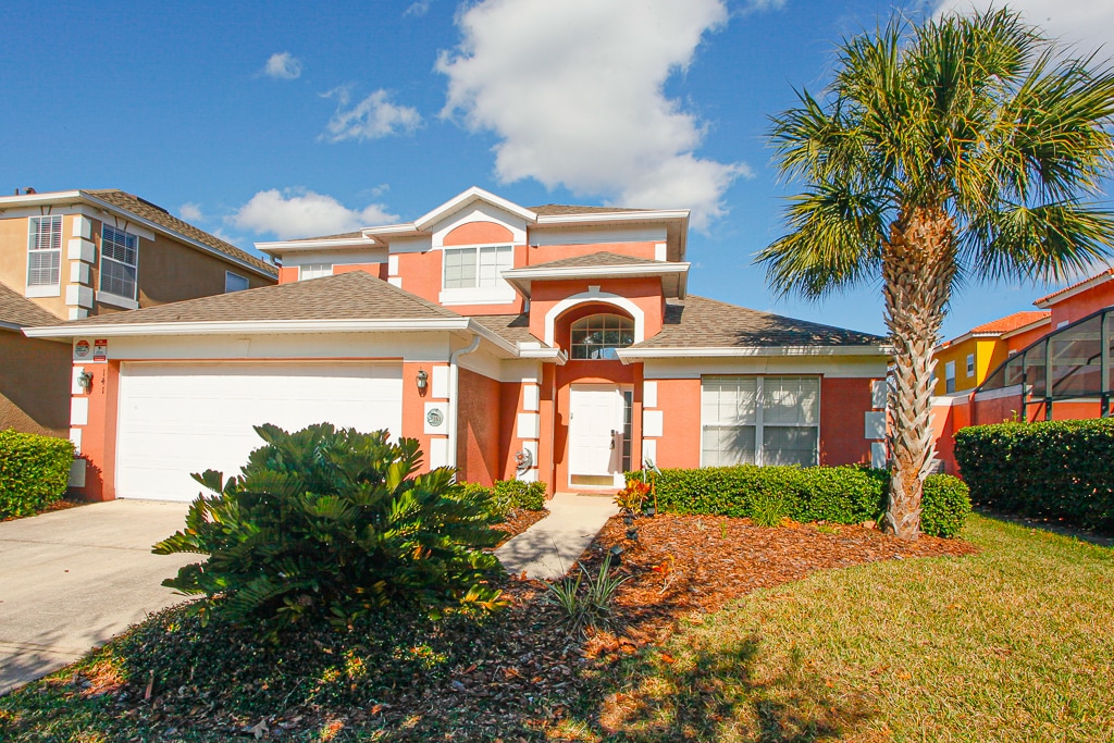 The exterior of the home is presented with a vibrant orange facade, framed by lush greenery and well-kept landscaping. Tall palm trees are featured prominently, and a welcoming pathway leads to the entrance, enhanced by large windows that provide ample natural light.