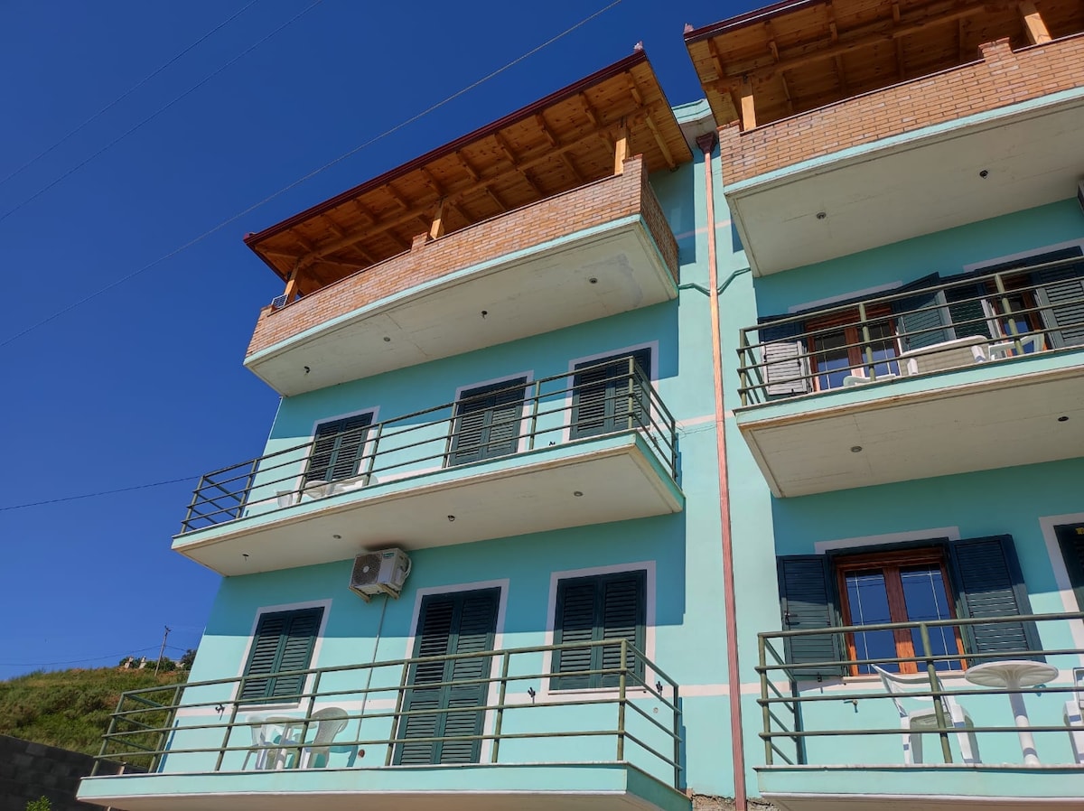 The exterior of the villa is shown, featuring a three-story structure painted in light blue. Multiple balconies with green railings are visible, with outdoor furniture placed on the second and third floors. Clear skies above complement the overall inviting appearance of the building.