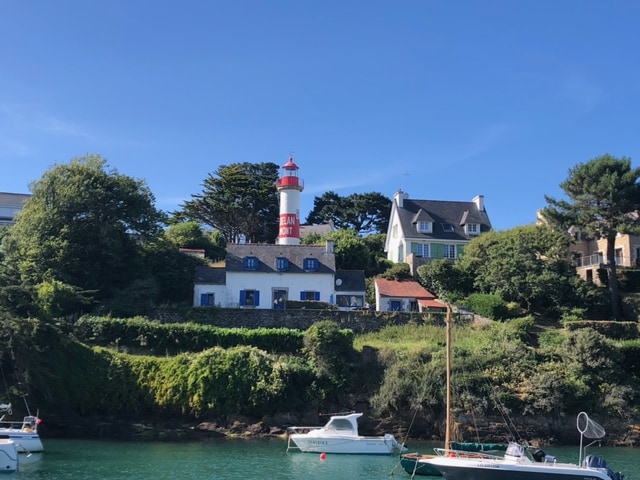 A coastal scene features a lighthouse with red and white stripes standing tall above a white building surrounded by lush greenery. The calm water reflects the natural surroundings, while several boats are moored nearby, providing a sense of maritime tranquility.