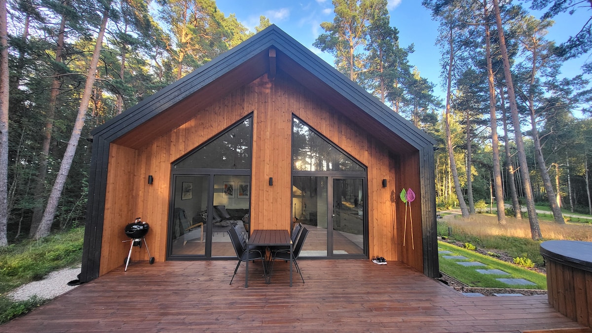 The exterior of a modern wooden cabin is framed by tall trees, featuring large glass doors that offer a view of the interior. A spacious deck includes a table and chairs, alongside a grill, enhancing the outdoor space.