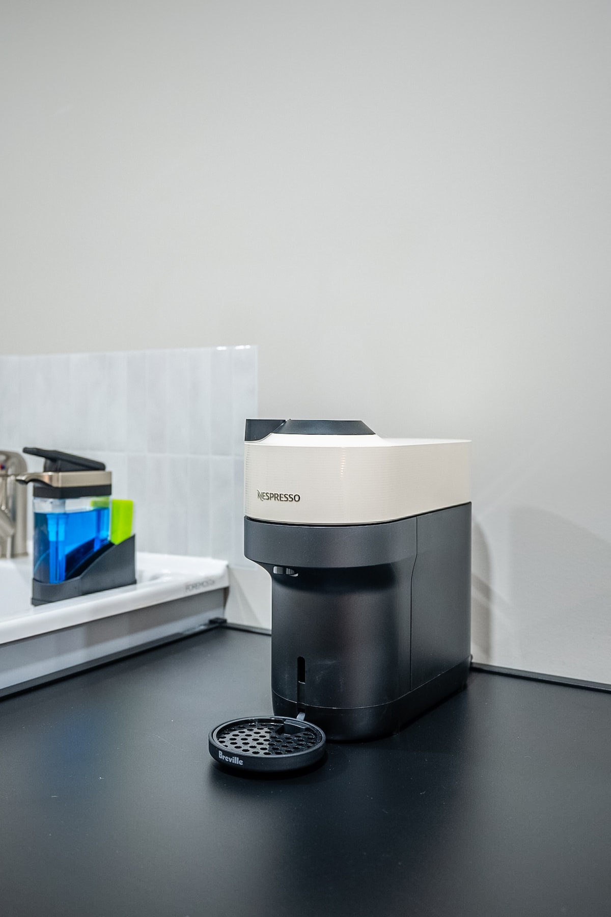 A modern Nespresso machine is positioned on a sleek black countertop, surrounded by a minimalist setup. A small dish sits next to the machine, designed for used capsules. A colorful cup rests on the counter, adding a touch of vibrancy to the space.