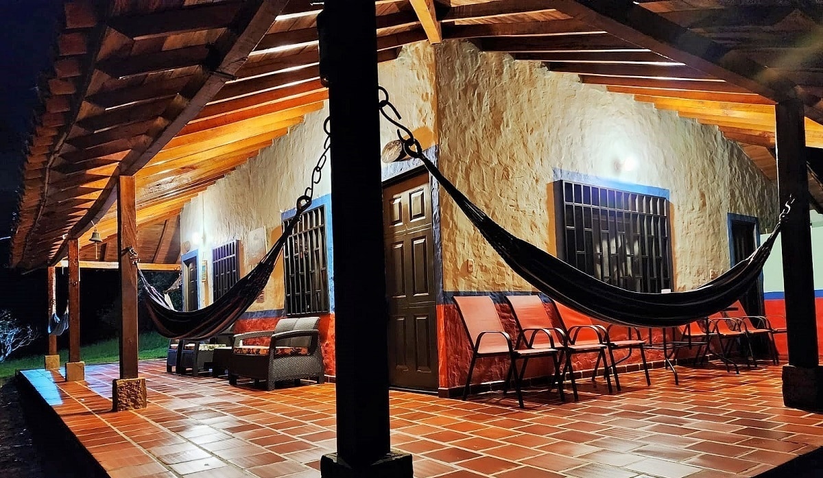 A spacious porch is seen with wooden beams overhead, featuring two hammocks and several chairs. The floor is tiled in warm hues, while light fixtures illuminate the area, creating a cozy atmosphere for relaxation. A main entrance door is visible, framed by windows.