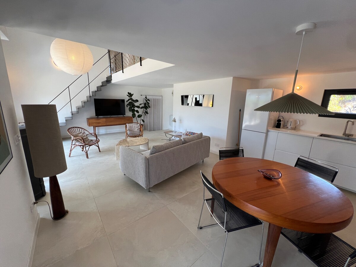 An open-concept living area is presented, featuring a light-colored sofa and a round wooden dining table with black chairs. A modern kitchen is visible in the background, and a staircase ascends to the upper level. Natural light fills the space, complemented by warm lighting fixtures.