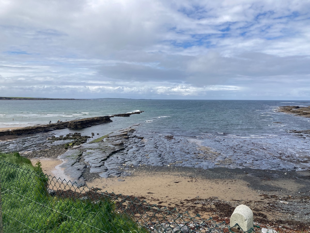 A scenic view of the coastline presents a mixture of rocky formations and sand, with calm waters gently lapping at the shore. The sky is partly cloudy, creating a serene atmosphere. A grassy area is visible in the foreground, framed by a low fence.