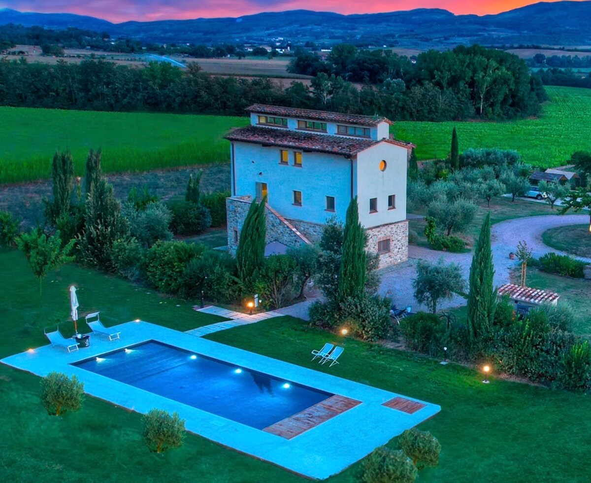 A spacious exterior view of the property showcases a charming stone building surrounded by lush greenery. A tranquil pool reflects the evening sky, with lounge chairs positioned nearby. The setting is enhanced by scenic farmland and gentle hills in the background.